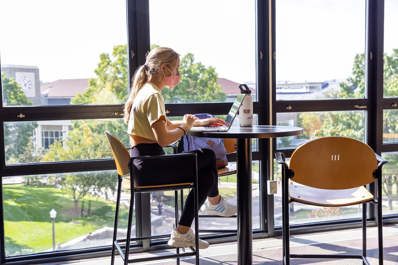 A student working on a laptop sits inside at a small round table near windows overlooking JMU's campus